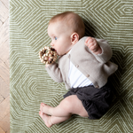 Little baby chews on wooden teether while being supported on a cushy floor mat designed for the whole family