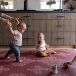 Sweet toddler boy walks across thick foam play mat by totter and tumble with a striking garnet red colour way and batik hand drawn motif that complements modern boho home furnishings another boy sits playing with toys as another adult clear toys out of shot