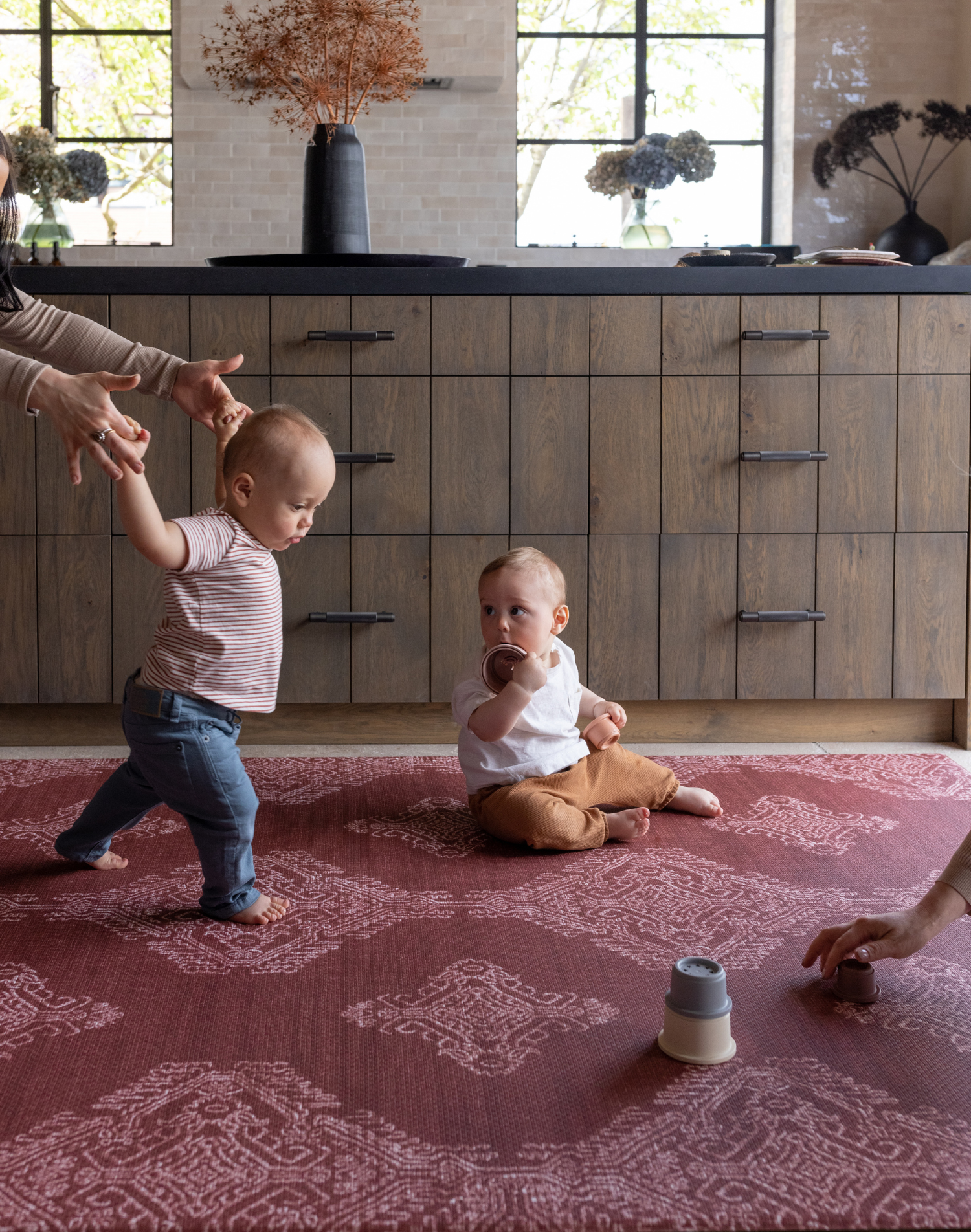 Sweet toddler boy walks across thick foam play mat by totter and tumble with a striking garnet red colour way and batik hand drawn motif that complements modern boho home furnishings another boy sits playing with toys as another adult clear toys out of shot