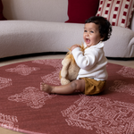 Baby girl sits on large round play mat with vibrant red design that is designed to complement modern family homes with a texture that allows it to sit like a rug for both style and protection