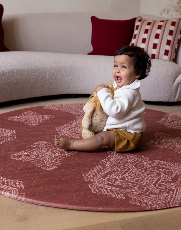 Baby girl sits on large round play mat with vibrant red design that is designed to complement modern family homes with a texture that allows it to sit like a rug for both style and protection