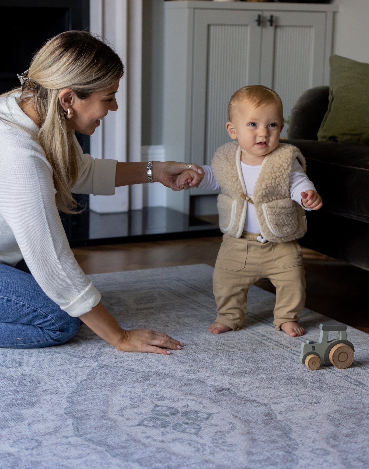 A mother holds the hand of a baby who is learning to walk. The baby is walking on a big soft foam mat that has a beautiful grey design